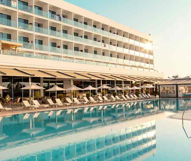 En hotellbyggnad med balkonger syns bakom en pool och solstolar. (AI-genererad text)