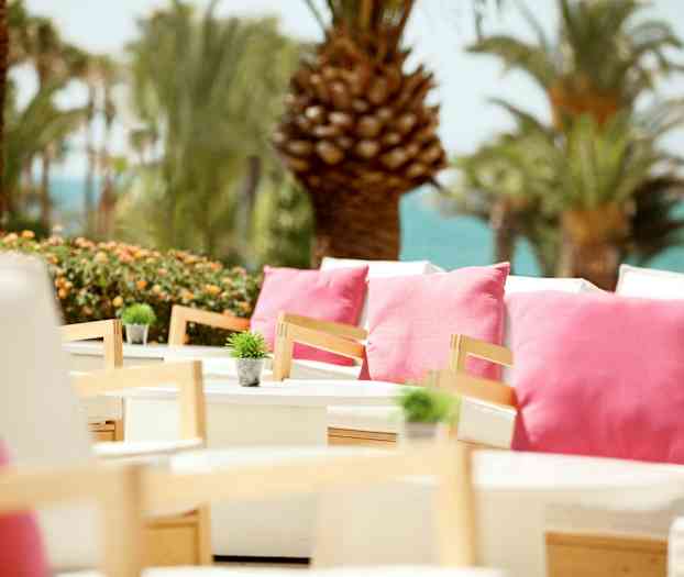 En utomhusterrass med vita sittmöbler dekorerade med rosa kuddar och små gröna växter, i en omgivning med palmer och en havsutsikt. (AI-genererad text)