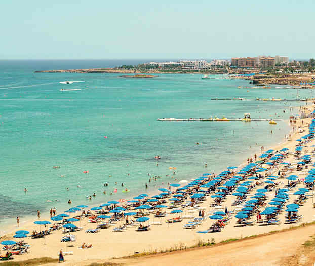 Utsikt över en strand med blå parasoller och badgäster vid det klarblå havet. (AI-genererad text)