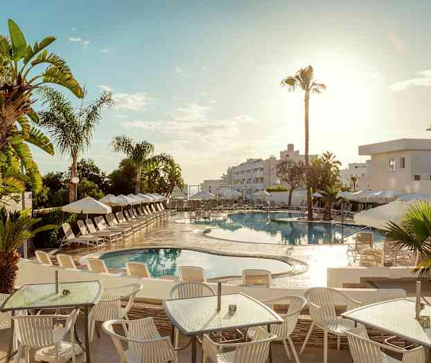 Bilden visar en utomhuspool på ett hotell, omgiven av solsängar och parasoller. I förgrunden finns bord och stolar under en solig himmel. (AI-genererad text)