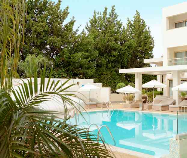 En utomhuspool omgiven av solstolar och parasoller vid ett hotell med grönskande träd i bakgrunden. (AI-genererad text)