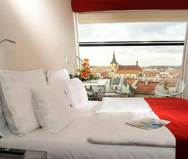 Ett hotellrum med en dubbelsäng, dekorerad med röda och vita sängkläder. En panoramautsikt över en stad med kyrkbyggnader syns genom fönstret. (AI-genererad text)