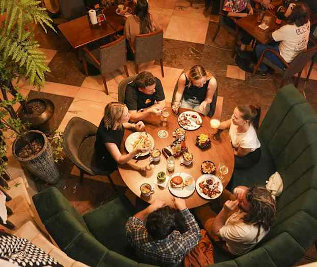 Fem personer sitter runt ett runt bord på en restaurang och äter och dricker. (AI-genererad text)