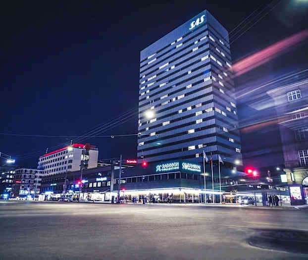 En hög byggnad med SAS-logotyp på toppen, Radisson Collection hotell på bottenvåningen, omgivande stadsbyggnader och nattmiljö. (AI-genererad text)