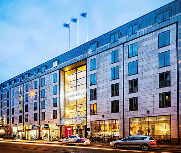 Exteriör av hotellbyggnad på kvällen, med belysning och gatan med passerande bilar. (AI-genererad text)
