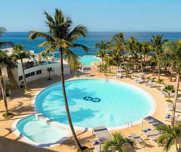 Ett hotellområde med en stor rund pool omgiven av palmer, solsängar och cabanas. Utsikt över havet i bakgrunden. (AI-genererad text)