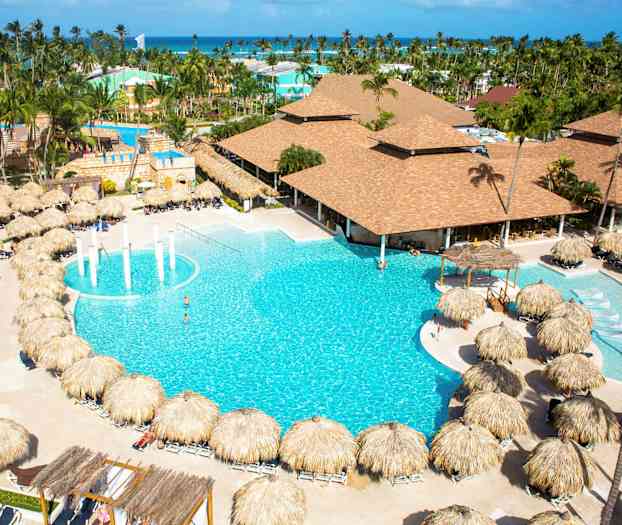 Ett hotellområde med en stor pool omgiven av solstolar med palmparasoller och byggnader i bakgrunden. (AI-genererad text)