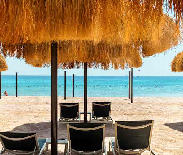 En sandstrand med solsängar och parasoller med halmtak, samt havet i bakgrunden. (AI-genererad text)