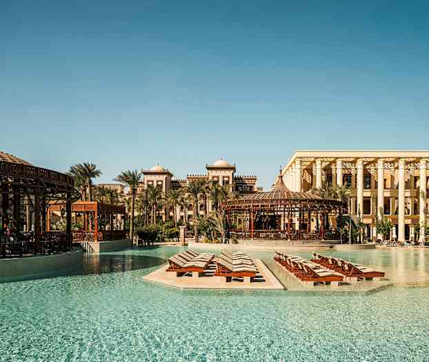 En vykortsliknande utomhusbild av ett lyxhotell med pool och liggstolar omgivna av palmer och arkitektur i traditionell stil. (AI-genererad text)