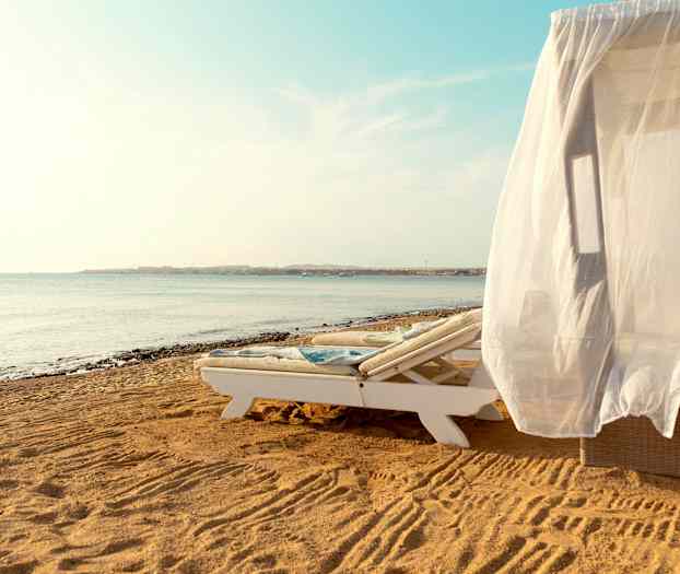 En solsäng med ett vitt draperi på en sandstrand vid havet. (AI-genererad text)