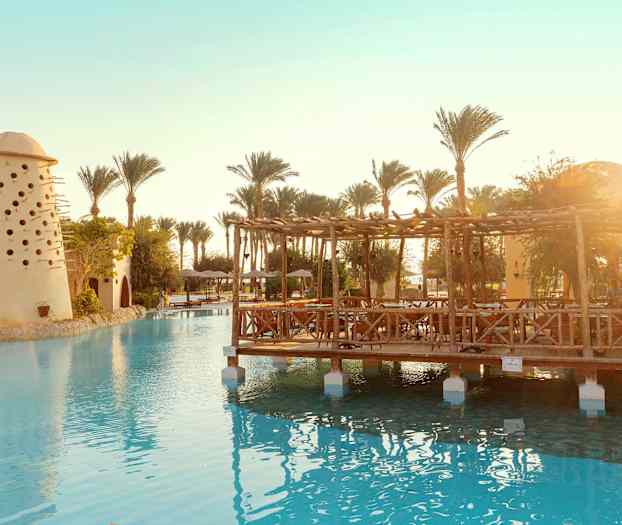 Bild av en swimmingpool med ett träpaviljong och en byggnad med små hål, omgiven av palmer. (AI-genererad text)