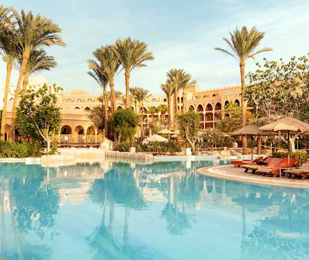 En stor pool omgiven av solstolar, parasoller, palmer och en byggnad i bakgrunden. (AI-genererad text)