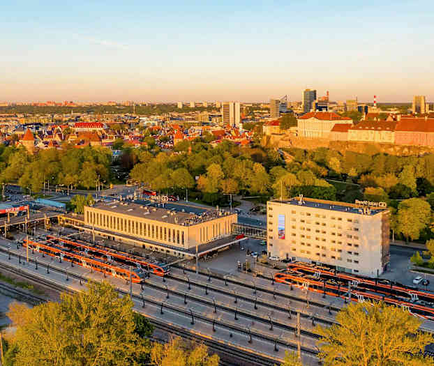 Bild av en stad med järnvägsstation i förgrunden och stadens silhuett i bakgrunden med byggnader och träd. (AI-genererad text)