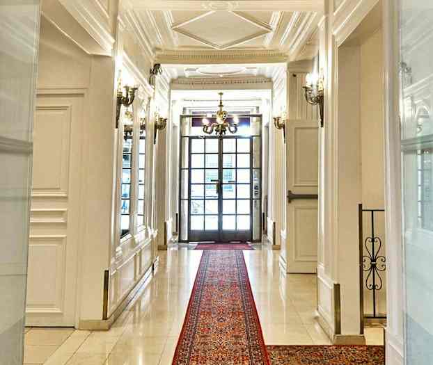 En elegant korridor med vita väggar och golv, en röd matta samt en ljuskrona och fönsterdörrar i bakgrunden. (AI-genererad text)