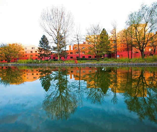 En bild av röda byggnader omgivna av träd, som speglas i en stilla vattenyta. (AI-genererad text)