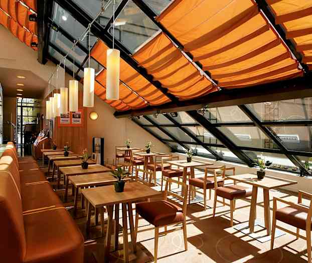 En ljus och modern matsal med träbord och stolar, orange takdekorationer och stora fönster. (AI-genererad text)