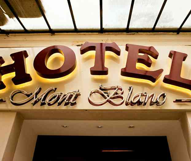 Ett hotellskylt med texten "HOTEL Mont Blanc", där "HOTEL" är i stora, belysta bokstäver och "Mont Blanc" är i en kursiv, dekorerad stil. (AI-genererad text)