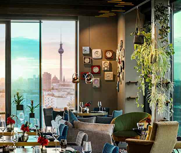 Ett elegant restauranginteriör med moderna möbler, gröna växter, och utsikt över en stadsbild med ett landmärke i bakgrunden. (AI-genererad text)