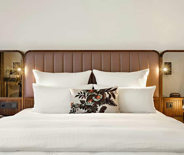 En dubbelsäng med vita lakan, flera kuddar och en dekorativ kudde med blommönster. Sänggaveln är brun med speglar på sidorna. (AI-genererad text)