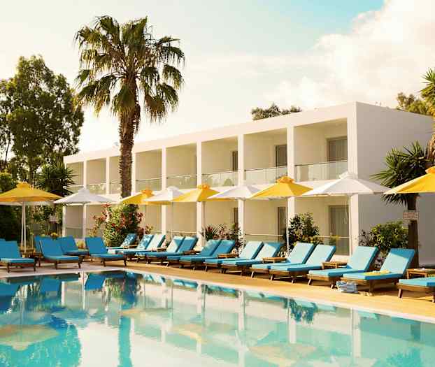 Ett hotell med vita byggnader och balkonger i en grön omgivning, framför en pool med solstolar och parasoller. (AI-genererad text)