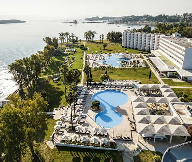 Flygvy över ett hotellområde vid havet med pooler, grönområden och byggnader. (AI-genererad text)