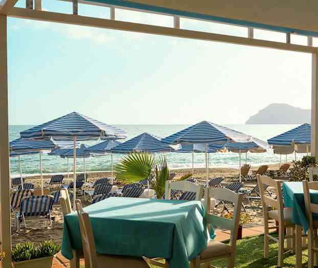 Strand med blå-vitrandiga parasoller och solstolar, bord i förgrunden. (AI-genererad text)