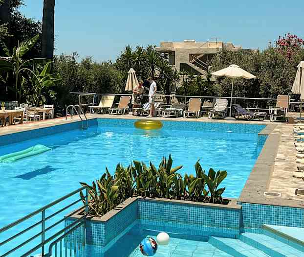 En swimmingpool med solsängar och parasoller runtomkring. I bakgrunden syns växtlighet och byggnader. (AI-genererad text)