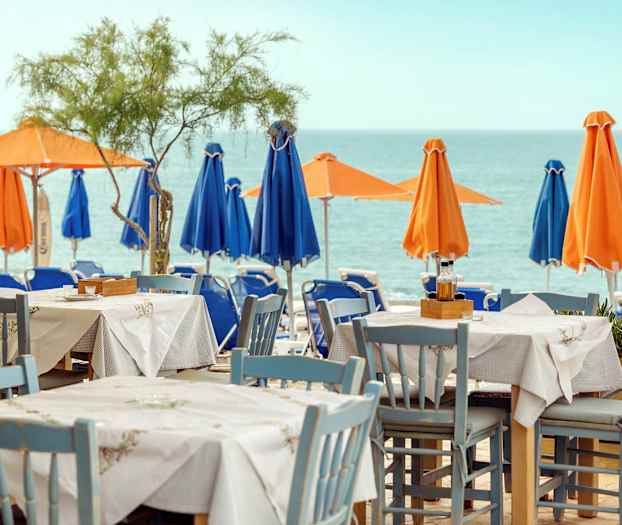 Ett bord och stolar står på en terrass med utsikt över havet. I bakgrunden syns parasoll i orange och blått på stranden. (AI-genererad text)