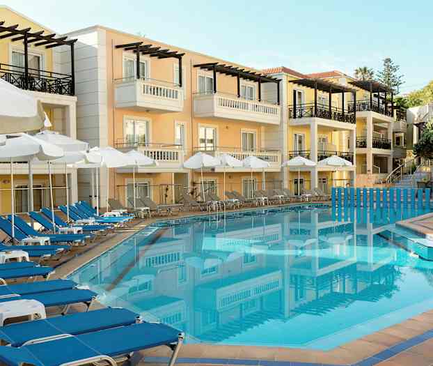 Hotellfasad i pastellfärger med balkonger, framför en klarblå swimmingpool omgiven av solstolar och parasoller. (AI-genererad text)