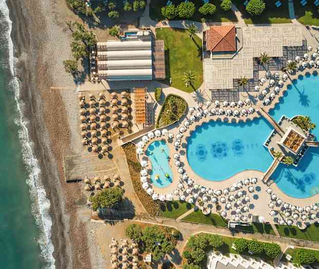 Flygvy över en strand med parasoll och solstolar bredvid ett stort poolområde med blå bassänger och vita parasoller. (AI-genererad text)
