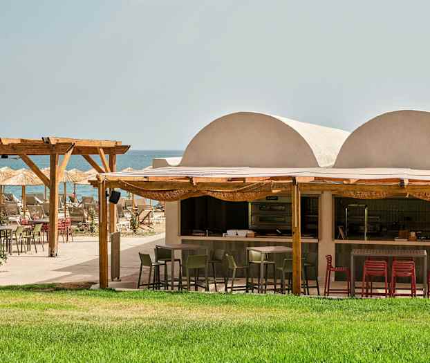 Utomhusrestaurang vid stranden med grön gräsmatta, träpergola och parasoller. (AI-genererad text)