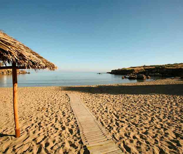 En sandstrand med utsikt över lugnt, klart vatten. Ett tak av vass syns till vänster. (AI-genererad text)