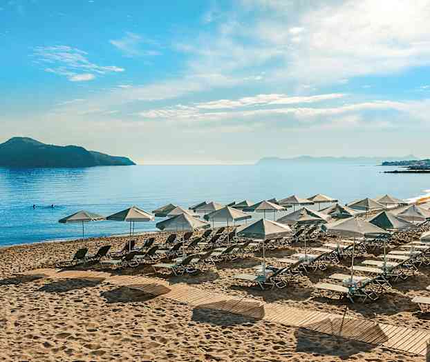 Solstolar och parasoller uppställda på en sandstrand vid en lugn havsutsikt med en ö och berg i bakgrunden. (AI-genererad text)