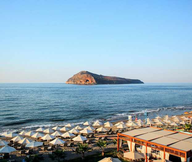 En strand med solstolar och parasoller vid havet, med en ö synlig i bakgrunden. (AI-genererad text)