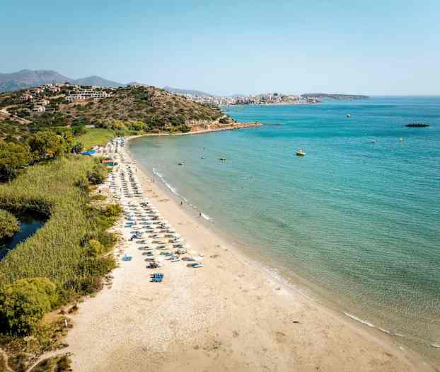 Sandstrand med solstolar vid havet, omgiven av grönska och berg i bakgrunden. (AI-genererad text)