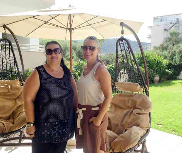 Två hänggungsstolar står under en stor parasoll på en terrass med utsikt över en trädgård och omgivande byggnader. (AI-genererad text)