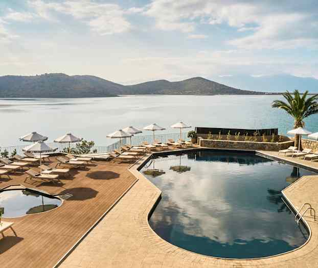 En pool med solstolar och parasoller vid havet, omgiven av berg och en palm. (AI-genererad text)