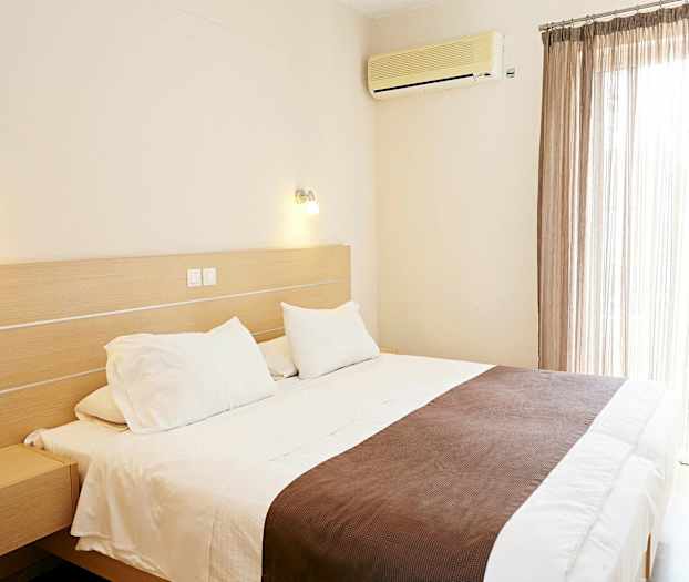 Ett hotellrum med en stor säng, vita sängkläder och brunt överkast. En luftkonditionering är monterad på väggen bredvid fönstret med ljusa gardiner. (AI-genererad text)
