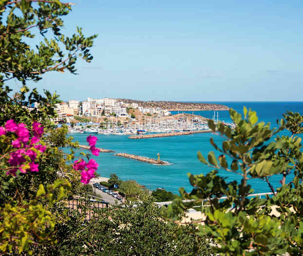 Bild av en hamn med segelbåtar omgiven av byggnader, sett genom grönska och rosa blommor. (AI-genererad text)