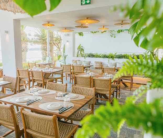 En restaurang med bord och stolar i trä, omgiven av grönska och med utsikt över havet. (AI-genererad text)