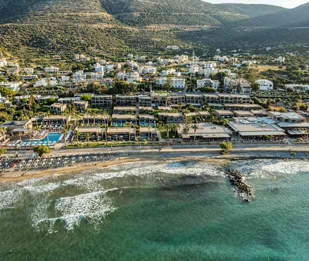 Vy över en strand med solstolar vid vattnet och hotellbyggnader i bakgrunden, med gröna kullar längre bort. (AI-genererad text)