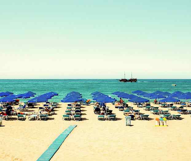 Strand med blå parasoller, solstolar och ett fartyg på havet. (AI-genererad text)