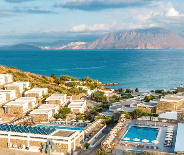 Bild av en kustnära resort med hotellbyggnader på sluttande mark omgivna av gröna områden och pooler, med hav och berg i bakgrunden. (AI-genererad text)