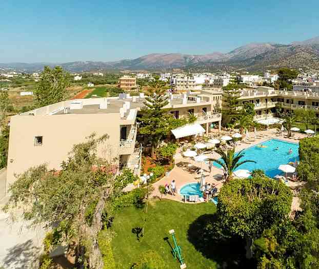 Luftvy av hotell med pool och trädgård, omgiven av grönska och byggnader med berg i bakgrunden. (AI-genererad text)