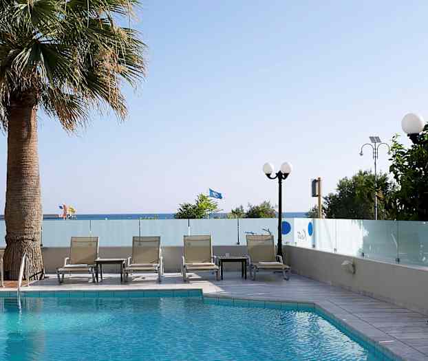 En pool med solstolar och ett bord vid sidan, omgiven av glasväggar. Träd och en vy mot havet i bakgrunden. (AI-genererad text)