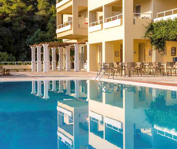 En utomhuspool framför en byggnad med sittplatser och grönska. (AI-genererad text)