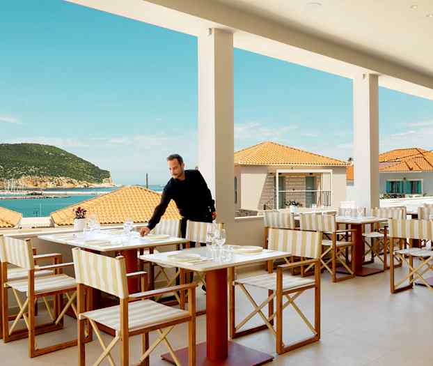 Restaurang med bordsuppsättning, utsikt över havet och terrass, mot bakgrund av terrakottatak och en grönskande bergskulle. (AI-genererad text)