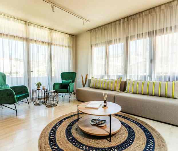 Ett ljust vardagsrum med stora fönster, två gröna fåtöljer, en beige soffa med randiga kuddar, och ett runt soffbord med dekorationer. (AI-genererad text)