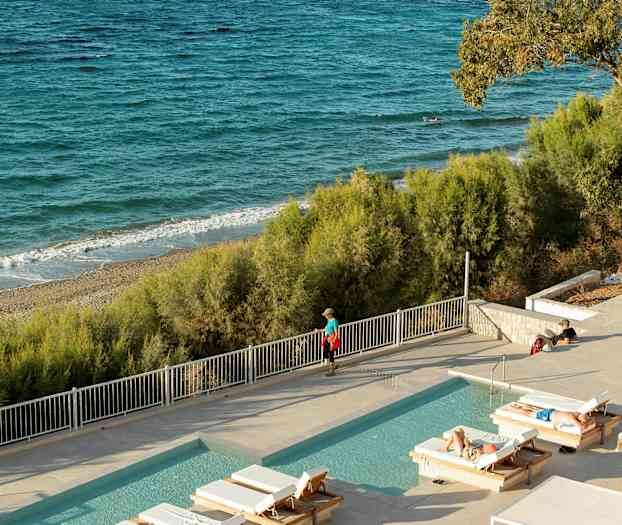 En utomhuspool med solstolar framför en sandstrand och havet. (AI-genererad text)