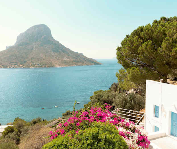 En vit byggnad med blå dörrar, omgiven av grönska och rosa blommor, med utsikt över havet och ett berg i bakgrunden. (AI-genererad text)